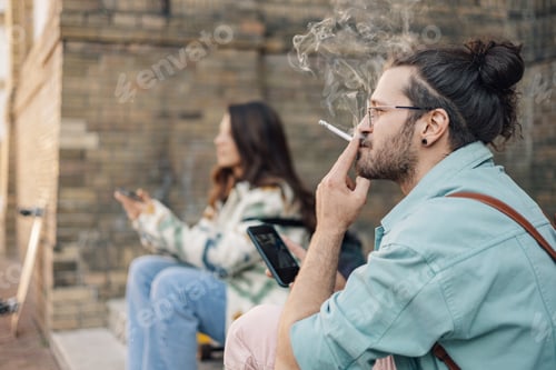 Preview: Side view of hipster guy using cellphone and smoking on street.