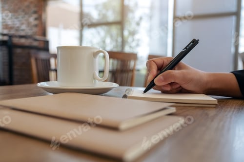 Preview: Closeup image of a hand writing on blank notebook with coffee cup on table in cafe