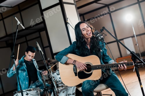 Preview: selective focus of beautiful inspired musician playing guitar and singing while mixed race man