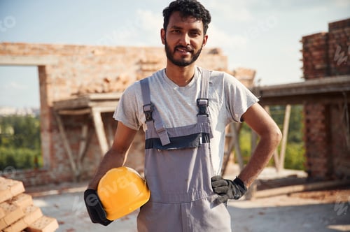 Preview: Standing and holding hard hat. Handsome Indian man is on the construction site