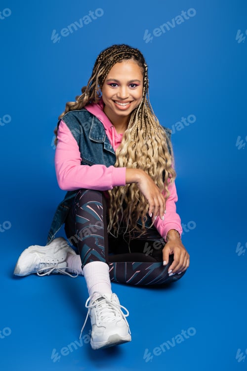 Preview: full length of happy long haired african american woman sitting in sporty outfit on blue background