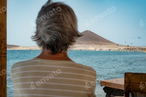 Preview: Rear view of mature woman in front of the sea looking at beautiful seascape