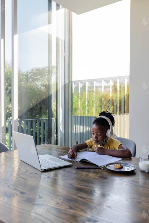 Preview: African American boy writing with red pencil at balcony table wearing headphones laptop, copy space