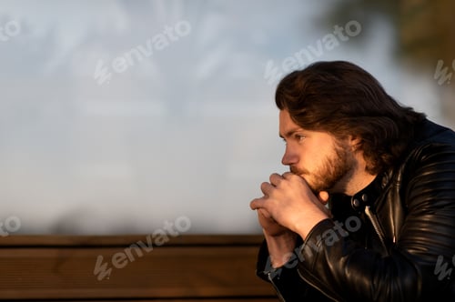 Preview: Pensive serious young bearded man wear black leather jacket sitting and thinking outdoors