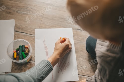 Preview: female hand draws on the floor on a white piece of paper with a colored pencil, child watching