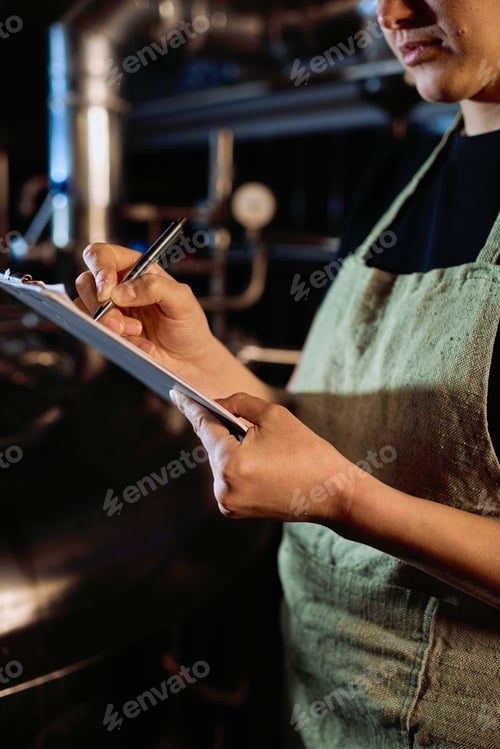 Preview: Young female brewer in apron pointing at paper while learning list of stages of beer preparation