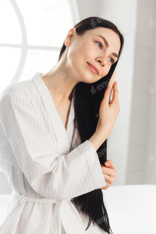 Preview: Young brunette woman combing her healthy and shiny hair sitting on bathtub weared in white bathrobe