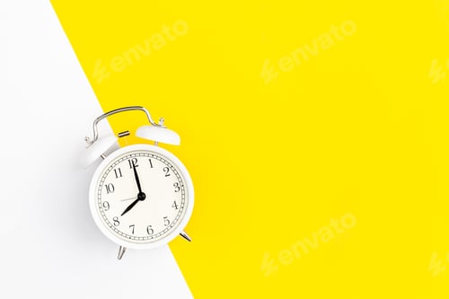 Preview: White alarm clock on a white and yellow background, flat lay.