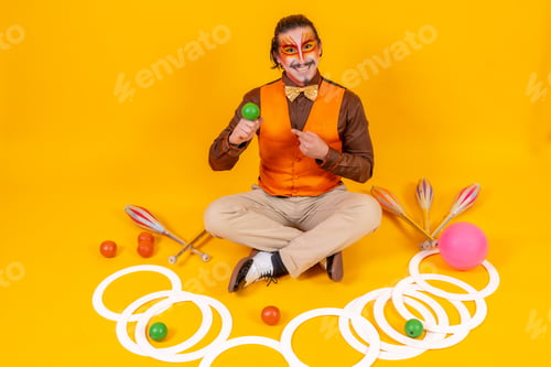 Preview: Portrait of a juggler in a vest and makeup sitting with the objects on a yellow background