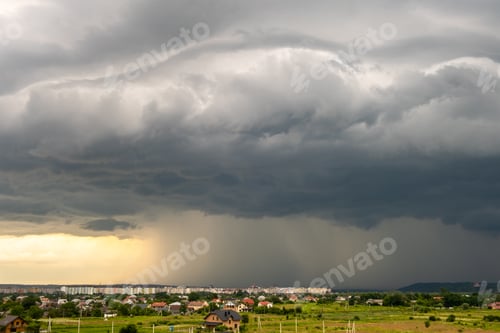 Preview: Moody landscape with dark stormy clouds with falling heavy downpour shower rain over distant town