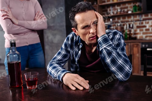 Preview: drunk man sitting at table while wife standing near by at home
