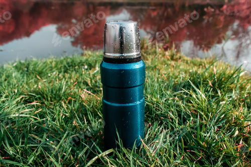 Preview: A camping thermos stands on the grass. autumn forest background. Vacuum metal thermos with raindrops