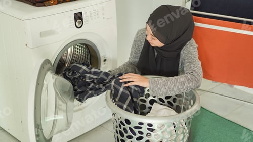 Preview: Asian woman in hijab puts dirty clothes in washing machine at home