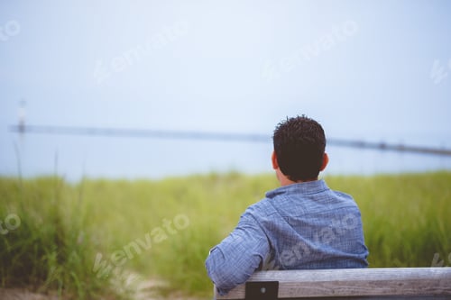 Preview: Male sitting on a wooden bench near the shoreline looking in the distance with blurred background
