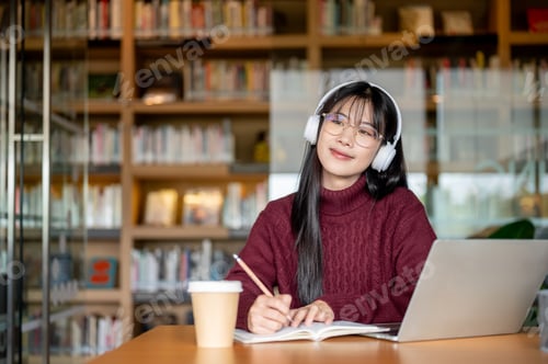 Preview: Asian woman wear headphones listening to music and writing in notebook aside laptop at library table