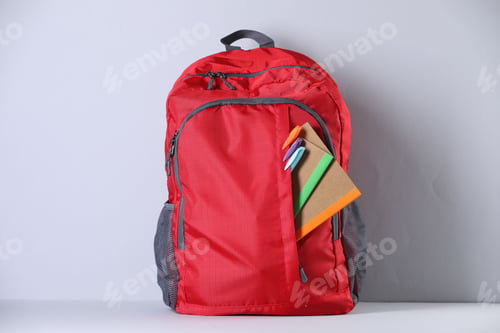 Preview: Red backpack with different stationery on table near light wall