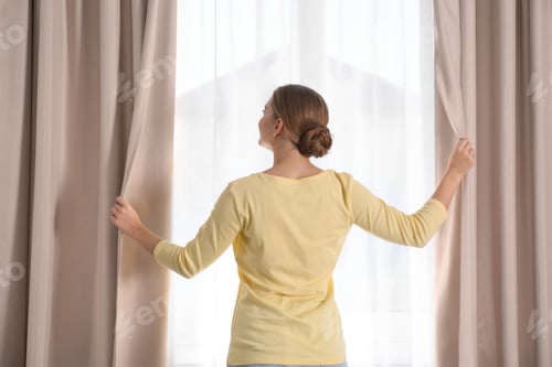 Preview: Woman opening window curtains at home in morning