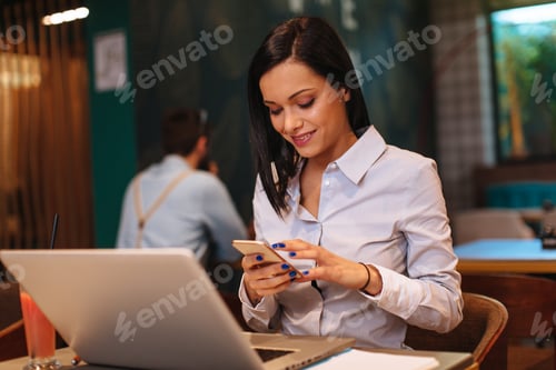 Preview: Young woman using smartphone in a cafe with a laptop in front of her