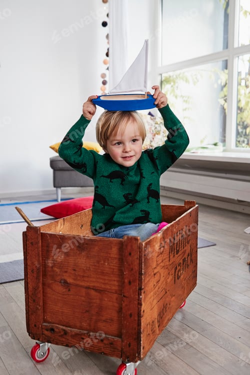 Preview: Young boy sitting in toy box, holding toy boat on head