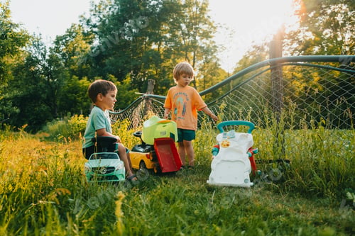 Preview: Cute little boys playing together on green summer garden background. Brothers riding plastic cars