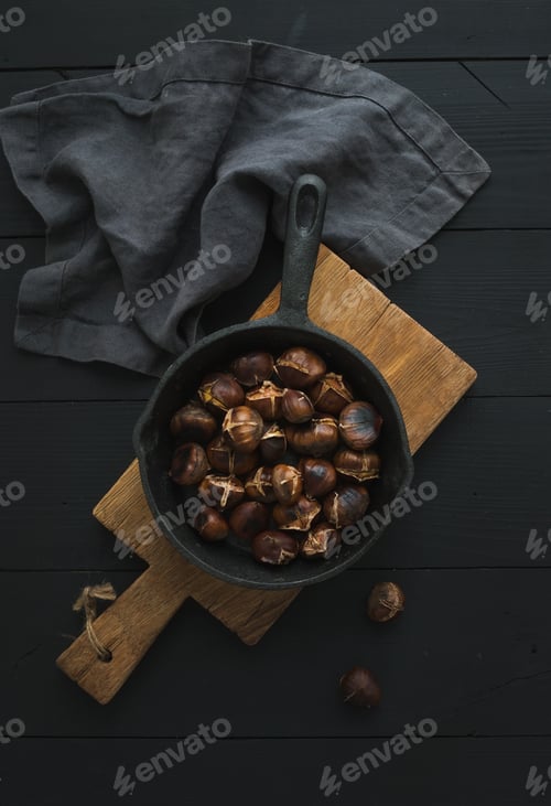 Preview: Roasted chestnuts in iron skillet pan on rustic wooden board over black table background, top view.