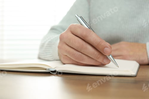 Preview: Man writing in notebook at wooden table, closeup