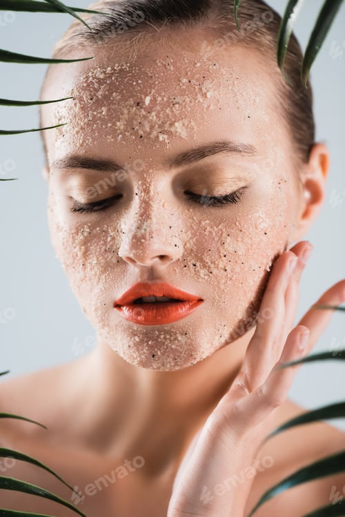 Preview: selective focus of naked woman with makeup and scrub touching face near palm leaves on white