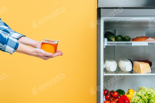 Preview: cropped view of woman holding yogurt near open fridge with fresh food on shelves isolated on yellow
