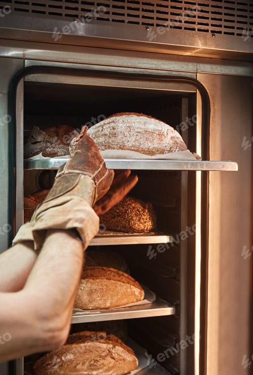Preview: Making a bread. Vertical photo of baker in working gloves taking the hot bread out of the oven