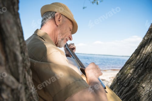Preview: Senior man in hat leaning on tree and playing guitar at riverside