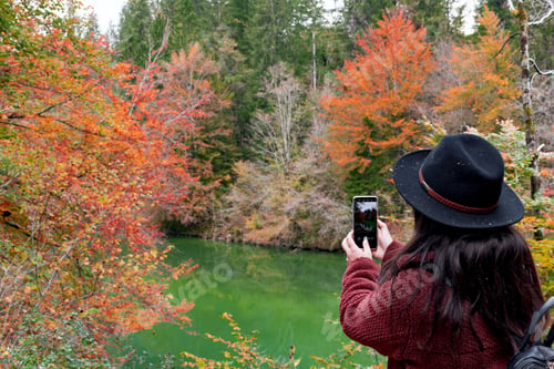 Preview: Rear view of woman wearing autumn clothes taking photo of amazing fall scenery with river and trees.