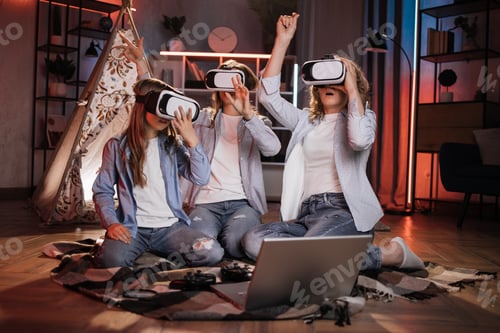 Preview: Happy mother and cute daughters sitting near toy wigwam at home and using modern VR headset