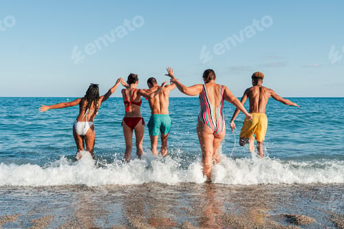 Preview: Rear view of a group of young people having fun on the beach, running to the sea to take a dip in