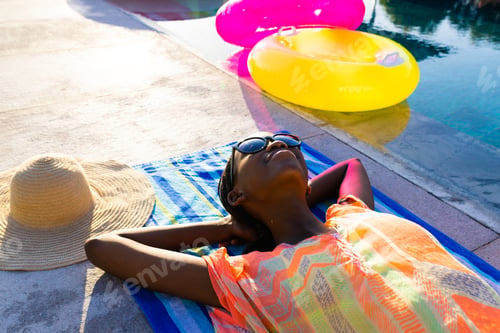 Preview: Happy african american woman lying on towel and sunbathing next to swimming pool in garden
