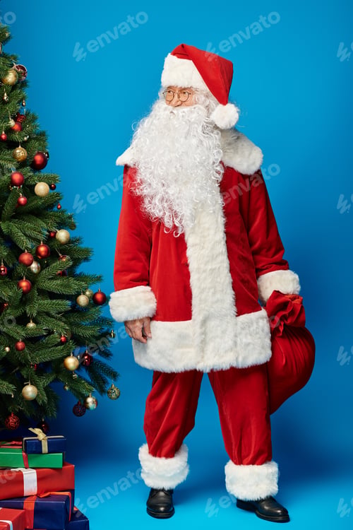 Preview: happy Santa Claus with beard and eyeglasses holding sack bag with presents near Christmas tree