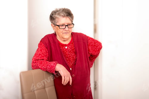 Preview: An old woman 70+ years old holds her hand behind her back, leans her elbows on a chair, looking tire
