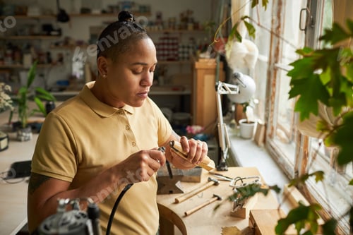Preview: Portrait of black female artist working on handmade jewelry in cozy workshop lit by sunlight