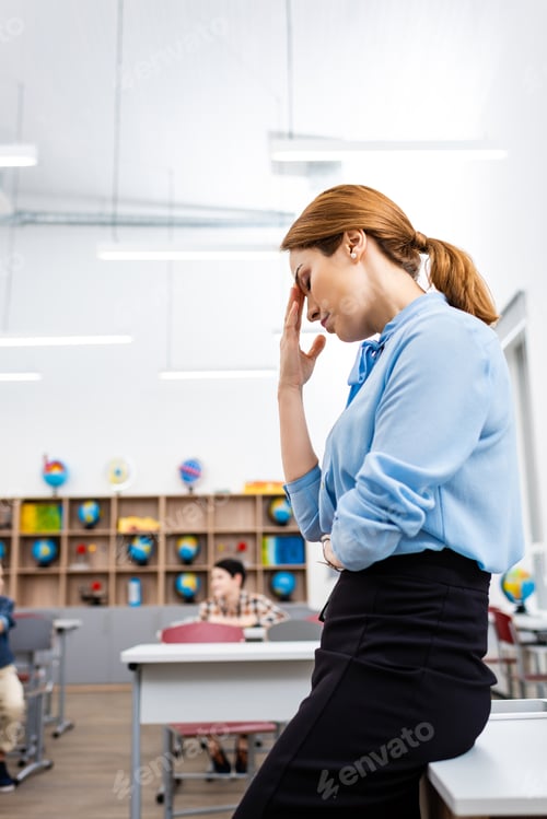 Preview: Tired teacher in blue blouse standing near table in classroom