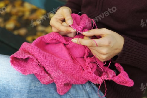 Preview: hands of woman knitting a pink sweater while sitting on park bench in autumn