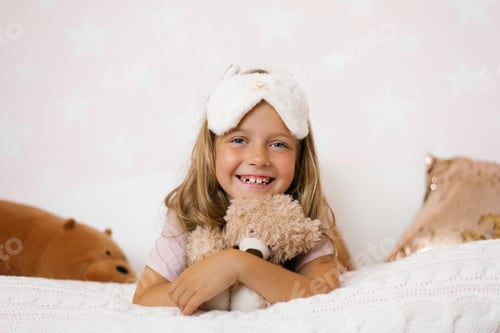 Preview: Portrait of a happy baby girl with a sleep mask on her head, lying on the bed hugging her soft toy
