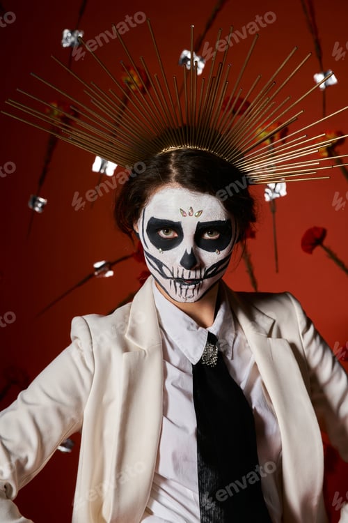 Preview: woman in dia de los muertos sugar skull makeup and crown looking at camera on red floral backdrop