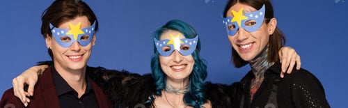 Preview: cheerful nonbinary person hugging queer friends in party masks and smiling at camera isolated on