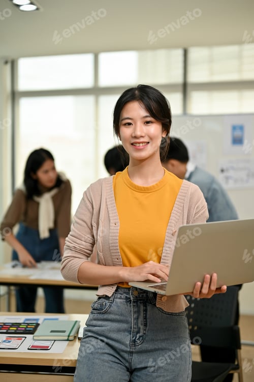Preview: A female graphic designer stands in the co-working office with a laptop in her hand.