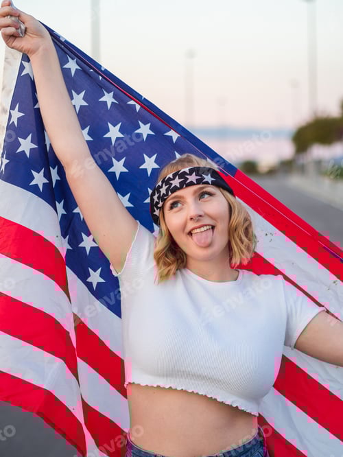 Preview: Vertical shot of a Caucasian woman from Spain holding the American flag behind her, tongue out