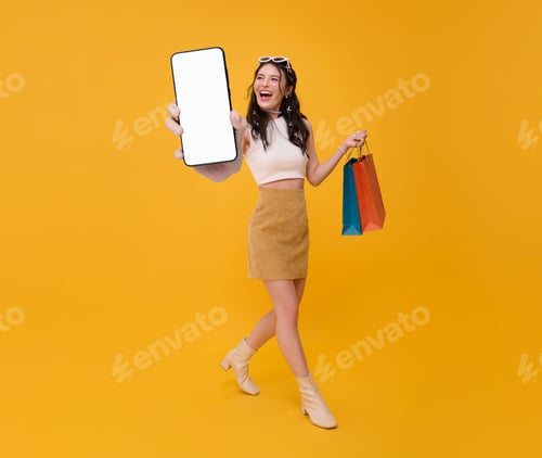 Preview: Happy Asian woman shopper showing smartphone blank screen and hand carrying shopping bags