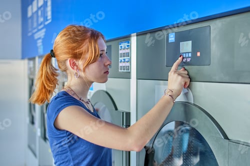 Preview: Young redhead woman with ponytail touching the buttons of a washing machine in a public laundry