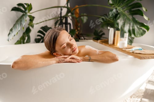 Preview: Dreamy young woman leaning on a bathtub side, relaxing with eyes closed at home