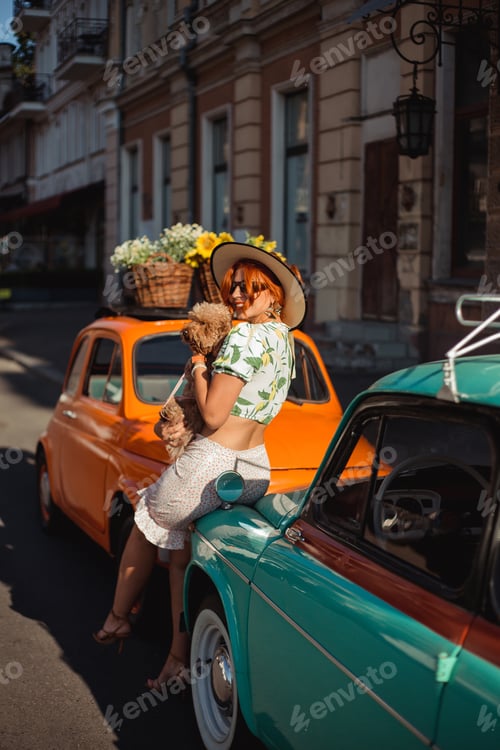 Preview: Young woman sitting on the hood of a retro car on a summer day playing with her puppy