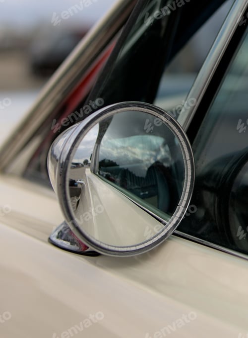 Preview: Vertical closeup of the round wing mirror of a vintage white carshowing the reflection of the road