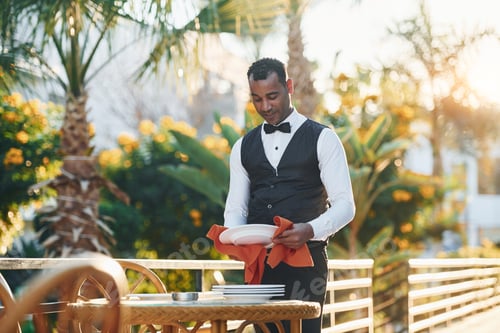 Preview: Wipes plates by red cloth. Black waiter in formal clothes is at his work outdoors at sunny daytime
